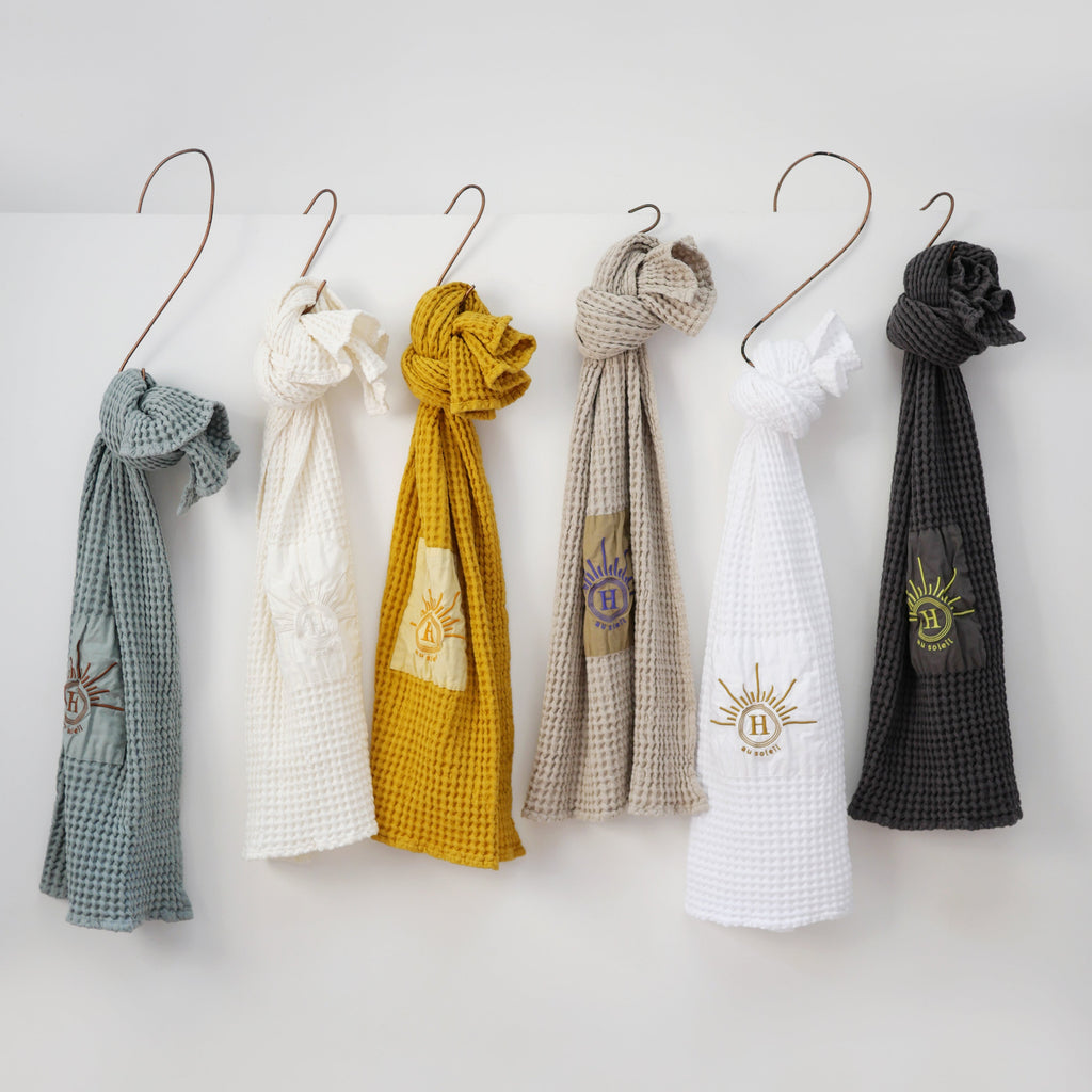 Collection de serviettes gaufrées en coton nid d’abeille de différentes couleurs – bleu, écru, moutarde, beige, blanc et gris – ornées de broderies soleil signées Honoré suspendu par crochet