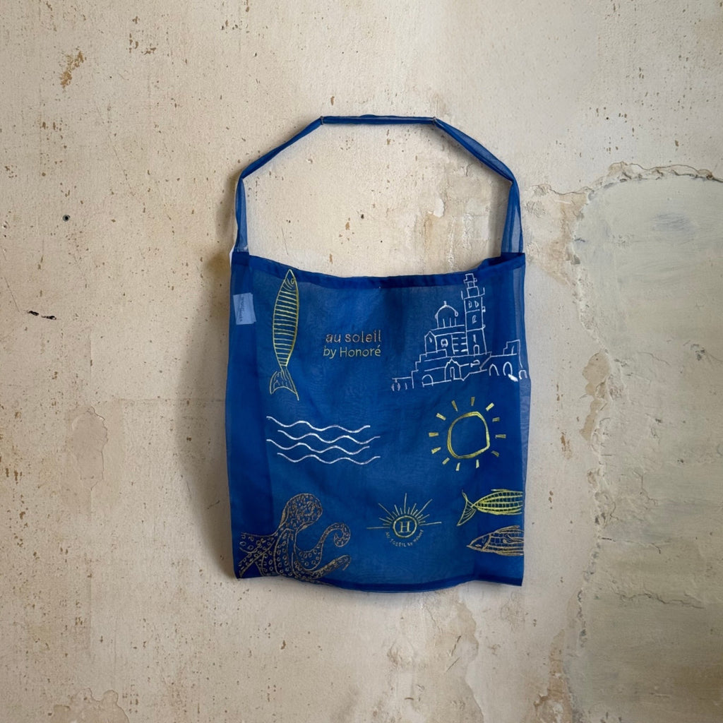 Sac tote bag en organza bleu indigo transparent, brodé de motifs inspirés de Marseille — le soleil, la mer, les poissons et la Bonne Mère — accompagné de l’inscription au soleil by Honoré. Une création légère et artisanale qui capture l’esprit méditerranéen et ensoleillé de la marque.