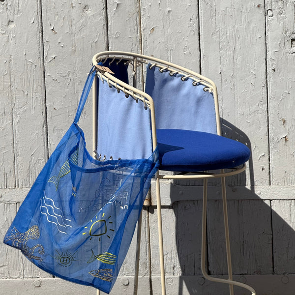 ac tote bag en organza bleu indigo transparent, brodé de motifs évoquant Marseille — le soleil, la mer, les poissons et la Bonne Mère. Suspendu à une chaise en métal blanc à assise bleue, il reflète l’esprit ensoleillé et méditerranéen d’Honoré, entre artisanat et légèreté.