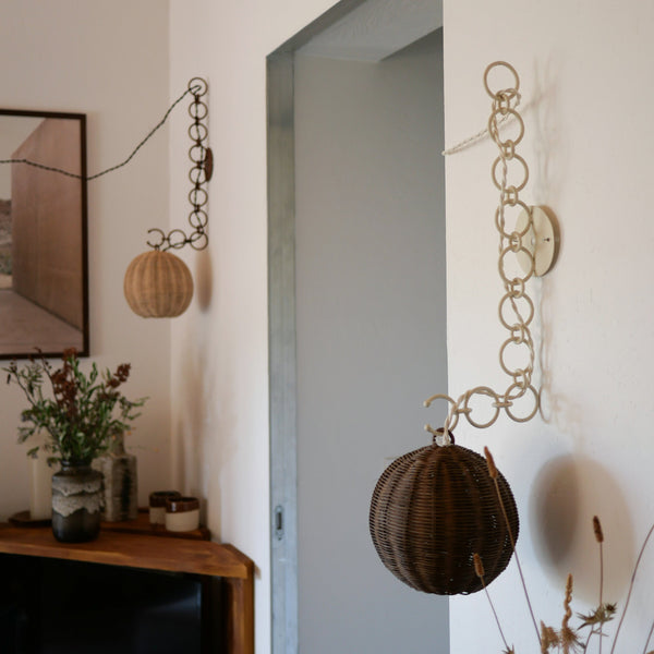Appliques murales en rotin suspendues à des chaînes décoratives en métal, installées de part et d’autre d’une ouverture. La scène dégage une ambiance naturelle et artisanale avec des matières brutes et des détails faits main, typiques du style méditerranéen d’intérieur.
