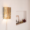 Applique murale en métal poli ajouré représentant un motif de soleil rayonnant, fixée sur un mur blanc. Son cordon torsadé en lin et la niche décorée d’objets en laiton créent une atmosphère chaleureuse et rétro, entre artisanat et design contemporain.