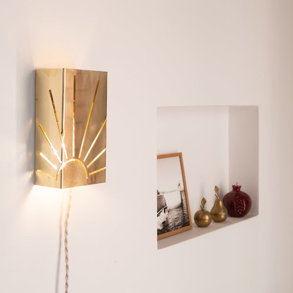 Applique murale en métal poli ajouré représentant un motif de soleil rayonnant, fixée sur un mur blanc. Son cordon torsadé en lin et la niche décorée d’objets en laiton créent une atmosphère chaleureuse et rétro, entre artisanat et design contemporain.