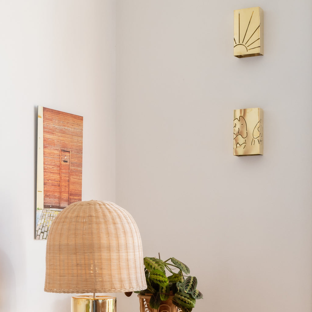Applique murale en métal doré poli ajouré représentant un motif de soleil et poisson rayonnant, fixée sur un mur blanc. Son cordon torsadé en lin et la niche décorée d’objets en laiton et de photos créent une atmosphère chaleureuse et rétro, entre artisanat et design contemporain, méditerannéen