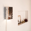 Applique murale en métal poli ajouré représentant un motif de soleil rayonnant, fixée sur un mur blanc. Son cordon torsadé en lin et la niche décorée d’objets en laiton et de photos créent une atmosphère chaleureuse et rétro, entre artisanat et design contemporain.