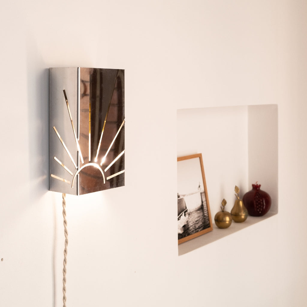 Applique murale en métal poli ajouré représentant un motif de soleil rayonnant, fixée sur un mur blanc. Son cordon torsadé en lin et la niche décorée d’objets en laiton et de photos créent une atmosphère chaleureuse et rétro, entre artisanat et design contemporain.