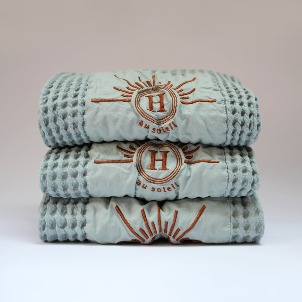 Ensemble de serviettes gaufrées en coton nid d’abeille couleur bleu céladon, avec broderie bronze emblématique Honoré en forme de soleil, empilées : linge de bain doux et raffiné taille 50x100 80x160