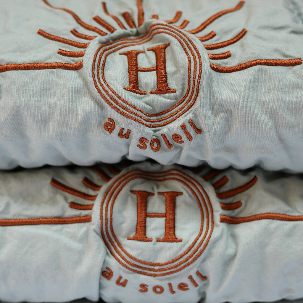 serviette de bain en coton nid d’abeille couleur bleu céladon, détail avec broderie bronze emblématique Honoré en forme de soleil, empilées : linge de bain doux et raffiné taille 50x100 80x160