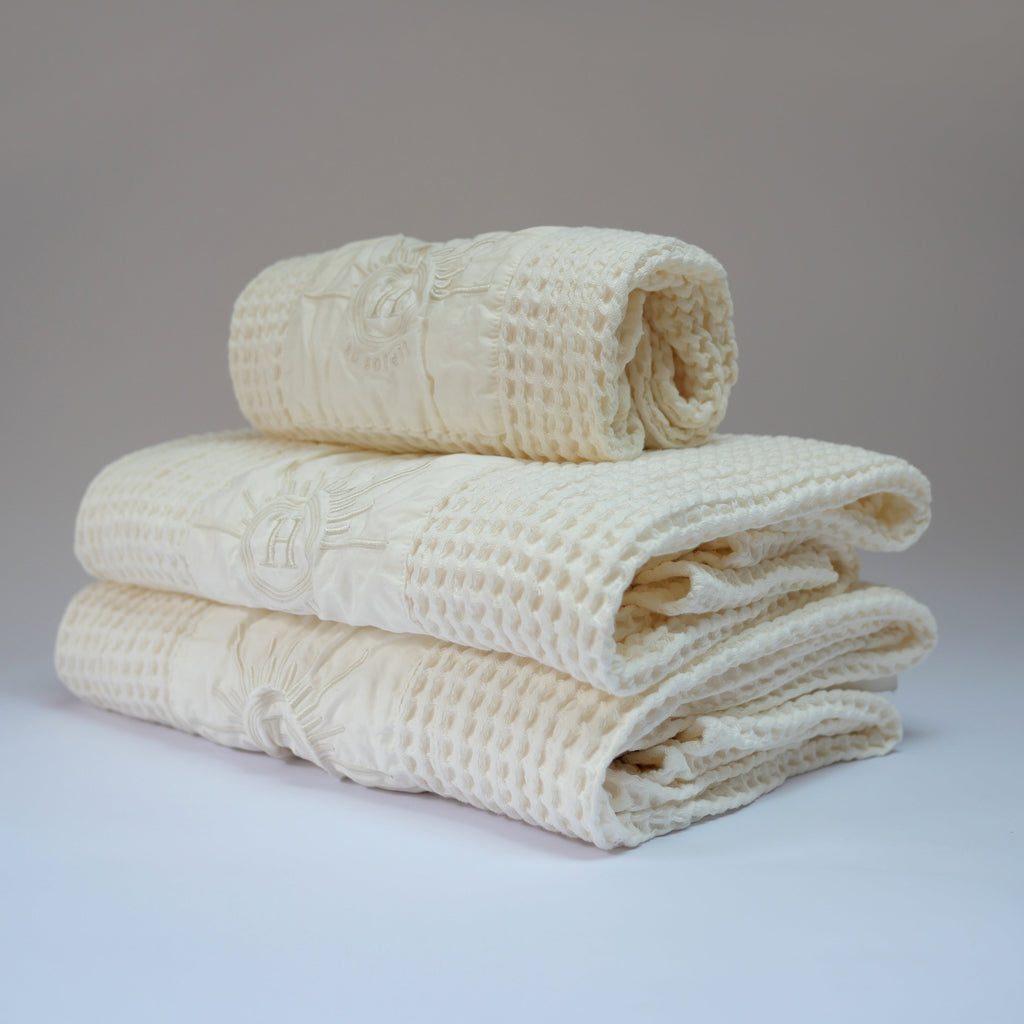 Ensemble de serviettes de bain gaufrées en coton nid d’abeille couleur écru, avec broderie emblématique Honoré en forme de soleil, empilées avec élégance, linge de bain doux et raffiné