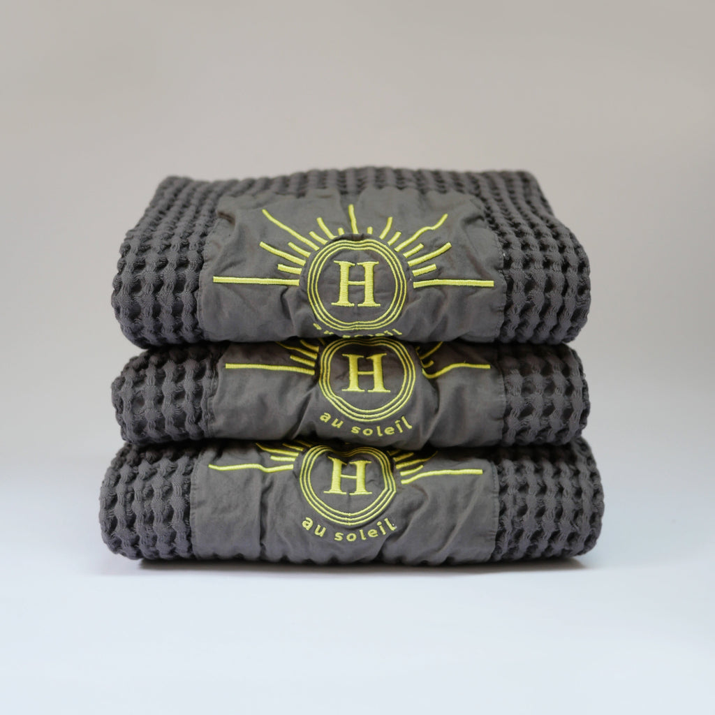 Ensemble de serviettes gaufrées en coton nid d’abeille couleur gris beton, avec broderie emblématique Honoré en forme de soleil, empilées avec élégance linge de bain doux et raffiné. 50 x 100 ou 80 x 160