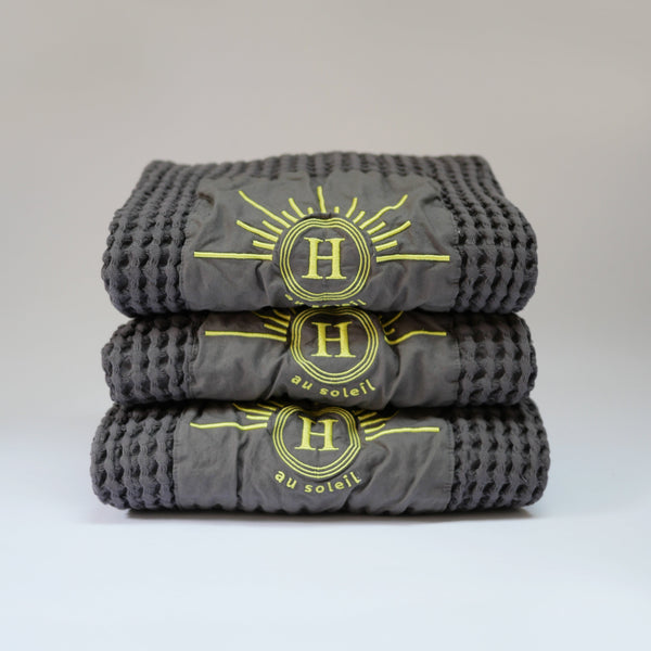 Ensemble de serviettes gaufrées en coton nid d’abeille couleur gris beton, avec broderie emblématique Honoré en forme de soleil, empilées avec élégance linge de bain doux et raffiné. 50 x 100 ou 80 x 160