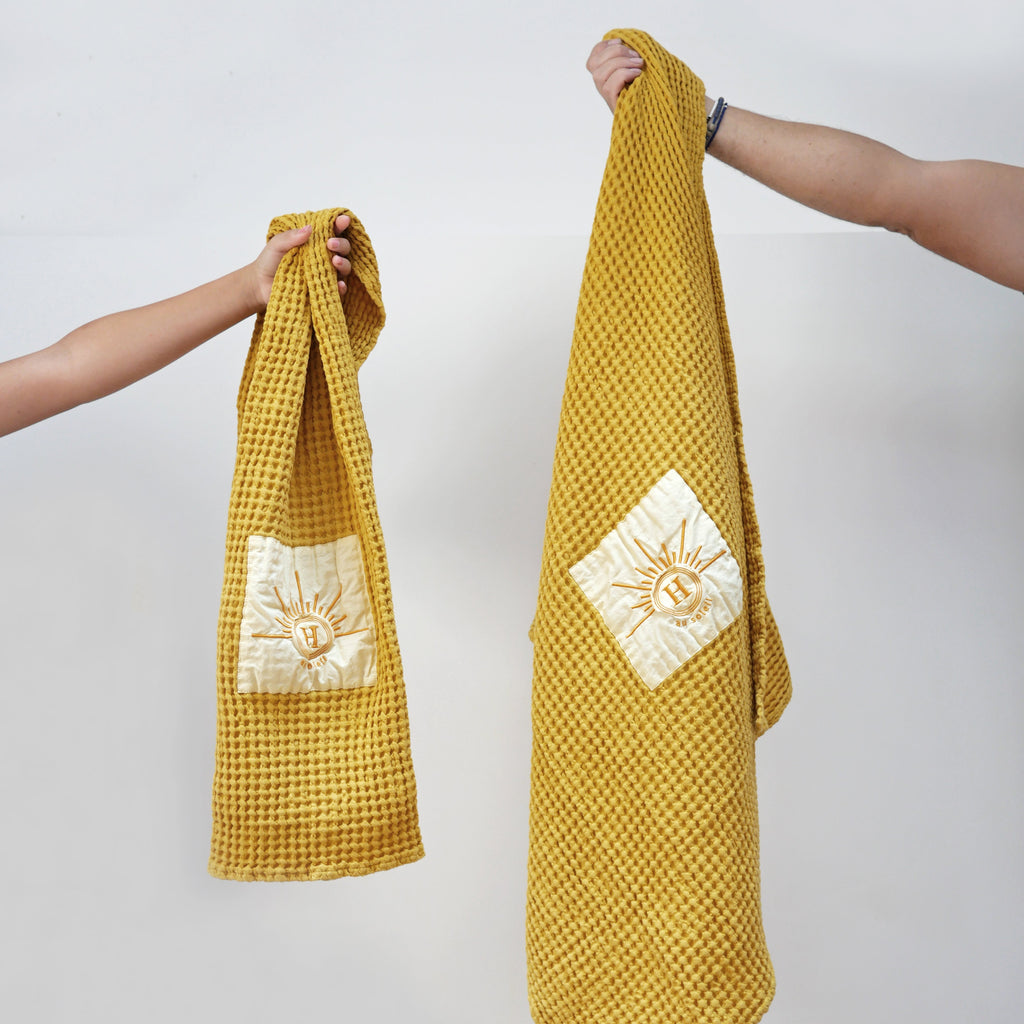 Serviettes gaufrées en coton nid d’abeille jaune, brodées du logo soleil Honoré, tenues à la main – linge de bain élégant et absorbant, au style artisanal.