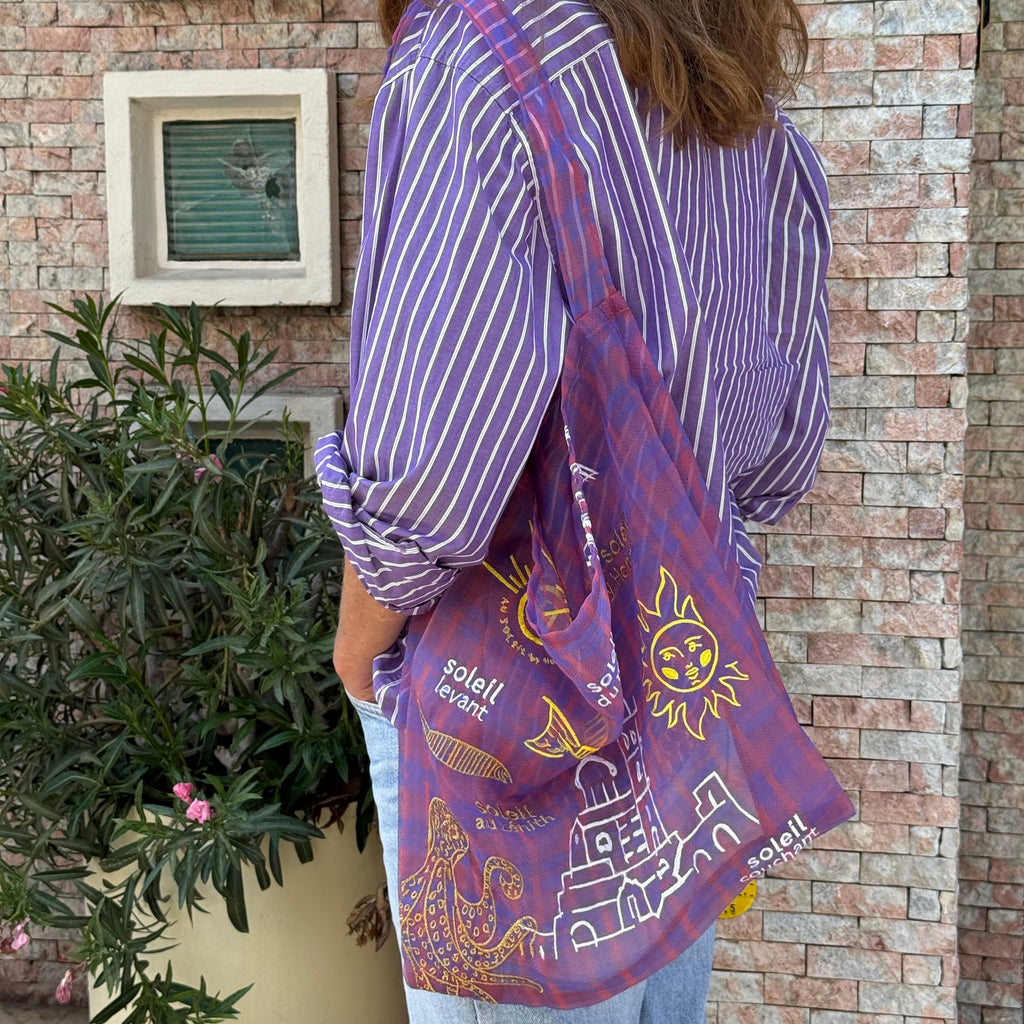 Sac type tote bag en organza imprimé tartan transparent, brodé de motifs évoquant Marseille — le soleil, la mer et la Bonne Mère — dans des tons chauds et lumineux. Porté à l’épaule sur une chemise rayée violette, il incarne un esprit méditerranéen, léger et artistique.