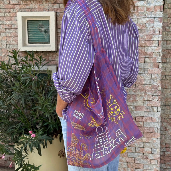 Sac type tote bag en organza imprimé tartan transparent, brodé de motifs évoquant Marseille — le soleil, la mer et la Bonne Mère — dans des tons chauds et lumineux. Porté à l’épaule sur une chemise rayée violette, il incarne un esprit méditerranéen, léger et artistique.