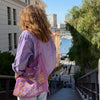 Sac type tote bag en organza imprimé tartan transparent, brodé de motifs évoquant Marseille — le soleil, la mer et la Bonne Mère — dans des tons chauds et lumineux. Porté à l’épaule sur une chemise rayée violette, il incarne un esprit méditerranéen, léger et artistique.