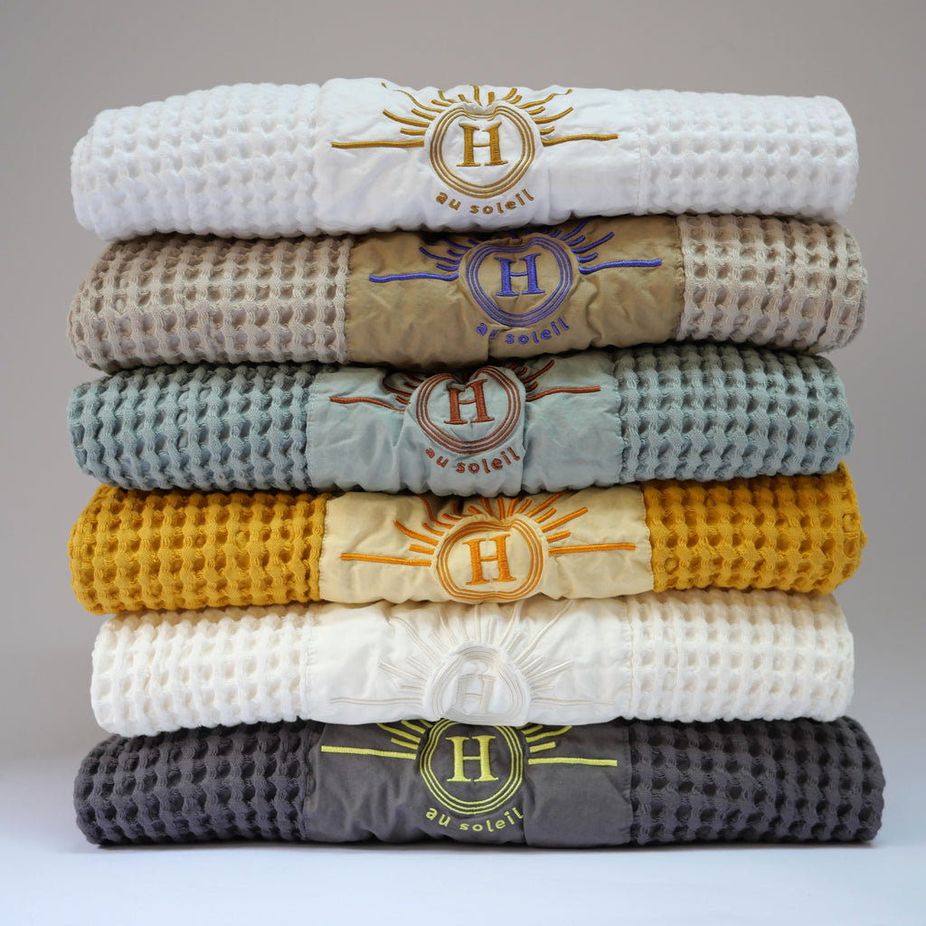 Collection de serviettes gaufrées en coton nid d’abeille de différentes couleurs – bleu, écru, moutarde, beige, blanc et gris – ornées de broderies soleil signées Honoré