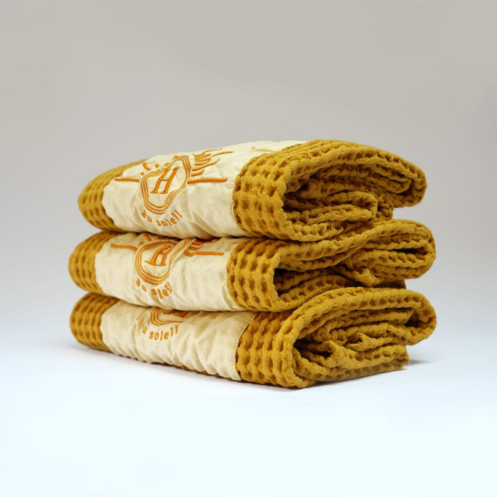 Ensemble de serviettes de bain gaufrées en coton nid d’abeille couleur jaune, avec broderie emblématique Honoré en forme de soleil, empilées avec élégance – linge de bain doux et raffiné. 50 x 100 cm ou 80 x 160 cm.