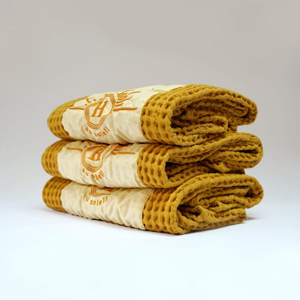 Ensemble de serviettes de bain gaufrées en coton nid d’abeille couleur jaune, avec broderie emblématique Honoré en forme de soleil, empilées avec élégance – linge de bain doux et raffiné. 50 x 100 cm ou 80 x 160 cm.