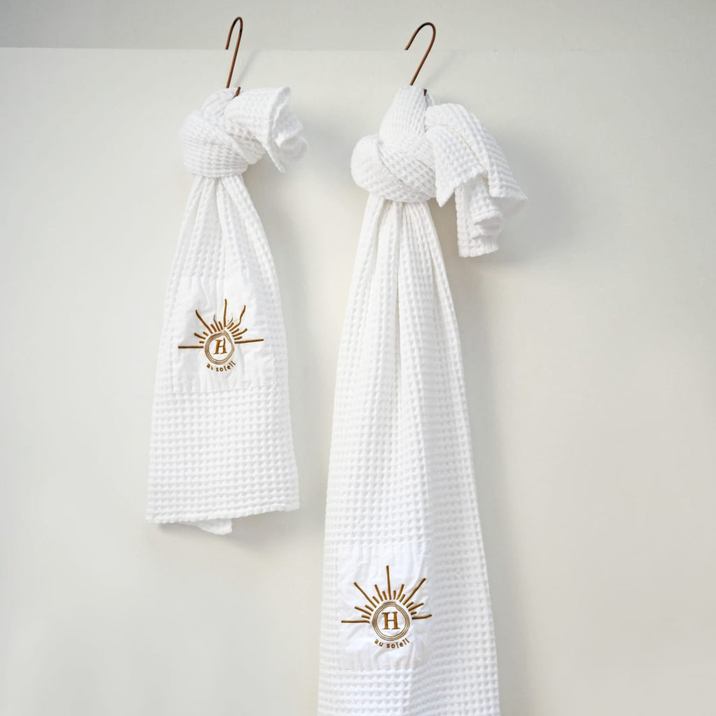 Collection de serviettes gaufrées en coton nid d’abeille blanc ornées de broderies soleil signées Honoré, suspendues sur des crochets décoratifs. Taille 50x100 80x160 