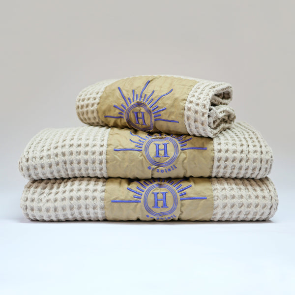 Ensemble de serviettes gaufrées en coton nid d’abeille couleur beige, avec broderie violet emblématique Honoré en forme de soleil, empilées avec élégance linge de bain doux et raffiné taille 50X100 80X160