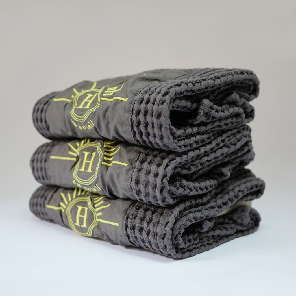 Ensemble de serviettes gaufrées en coton nid d’abeille couleur béton, avec broderie emblématique Honoré en forme de soleil, empilées avec élégance – linge de bain doux et raffiné.