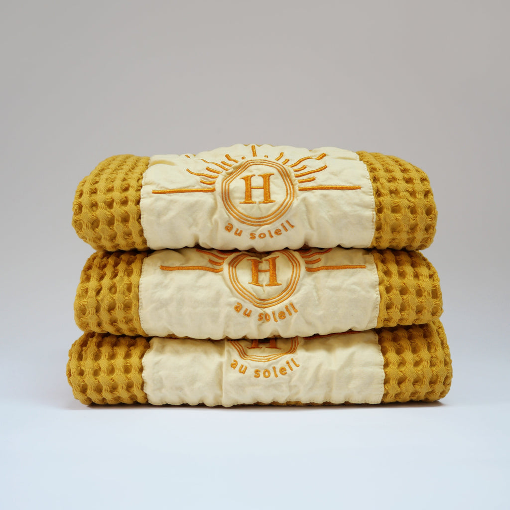 Ensemble de serviettes gaufrées en coton nid d’abeille couleur jaune, avec broderie emblématique Honoré en forme de soleil, empilées avec élégance, linge de bain doux et raffiné