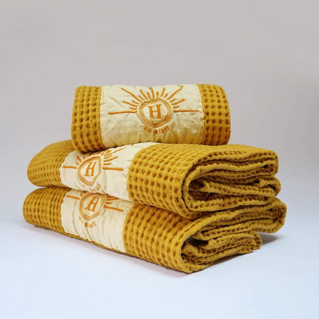 Ensemble de serviettes de bain gaufrées en coton nid d’abeille couleur jaune, avec broderie emblématique Honoré en forme de soleil, empilées avec élégance – linge de bain doux et raffiné.50 x 100 cm ou 80 x 160 cm