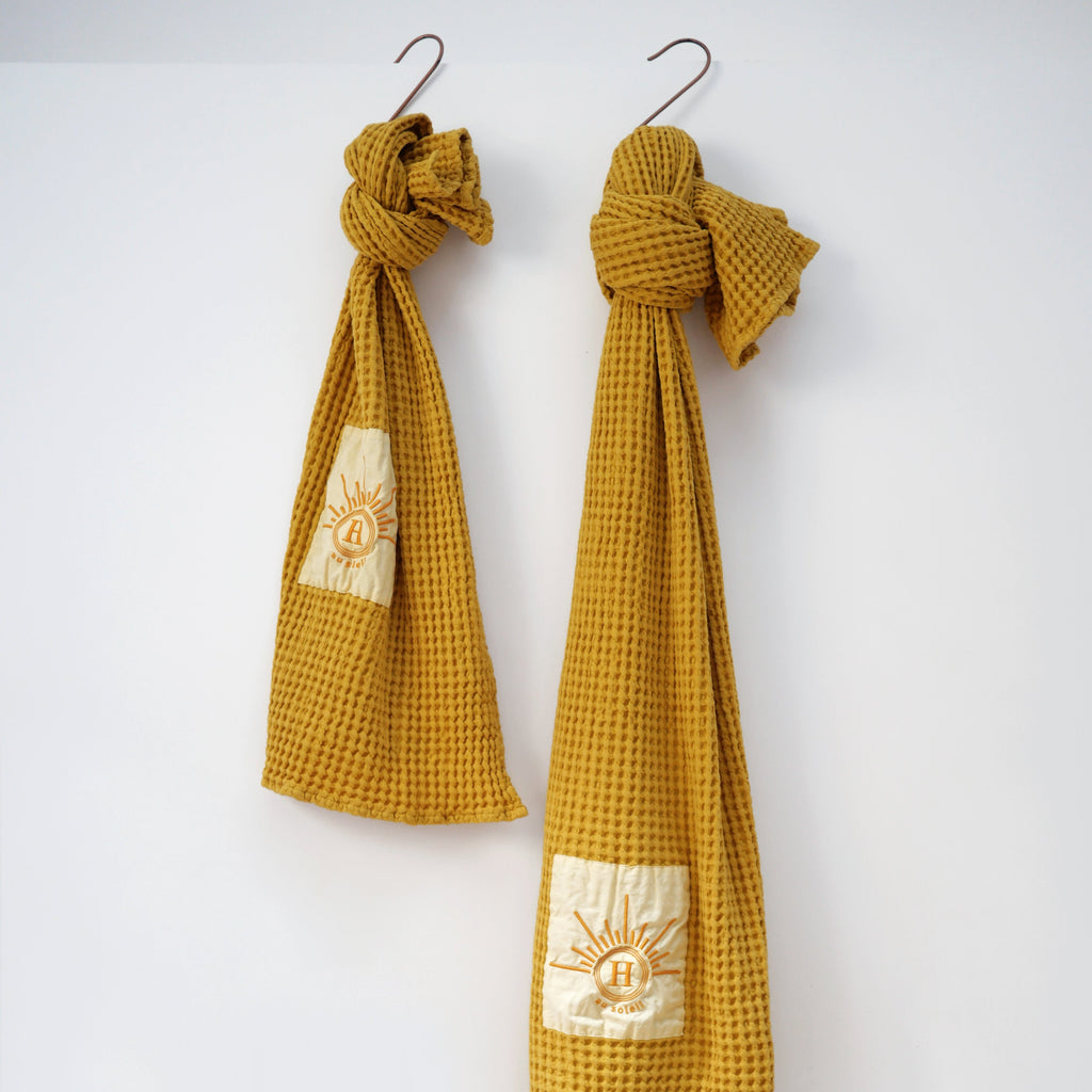 Collection de serviettes gaufrées en coton nid d’abeille moutarde ornées de broderies soleil signées Honoré, suspendues sur des crochets décoratifs.