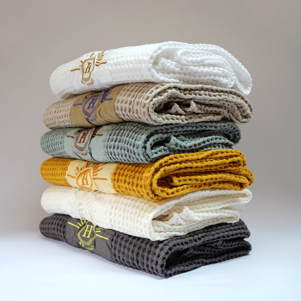 Collection de serviettes gaufrées en coton nid d’abeille de différentes couleurs – bleu, écru, moutarde, beige, blanc et gris – ornées de broderies soleil signées Honoré