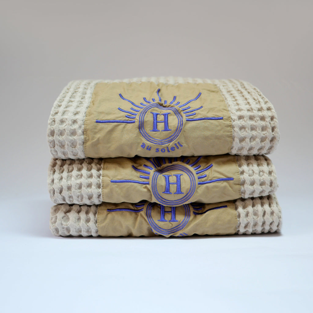 Ensemble de serviettes gaufrées en coton nid d’abeille couleur beige, avec broderie violet emblématique Honoré en forme de soleil, empilées avec élégance linge de bain doux et raffiné taille 50X100 80X160